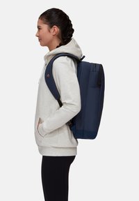 Marinblå ryggsäck med vadderade axelremmar och flera dragkedjefack. Slät textur med en strukturerad form, bärs över en ljus hoodie.