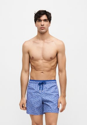 Jeune homme debout, torse nu, portant un short de bain bleu à motifs avec un cordon, sur fond blanc uni.