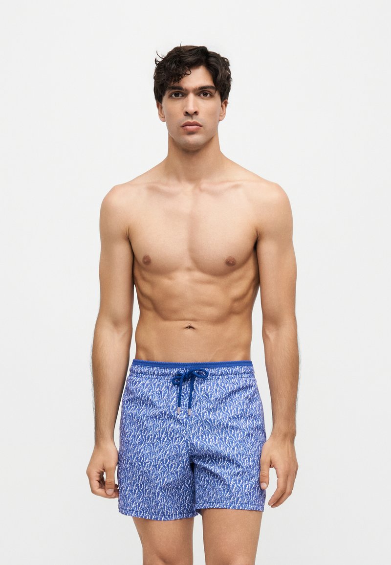 Jeune homme debout, torse nu, portant un short de bain bleu à motifs avec un cordon, sur fond blanc uni.