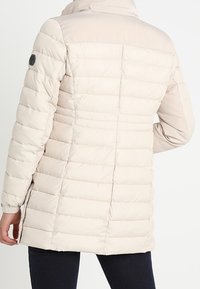 Beige pufferjakke med en tætsiddende silhuet, quiltet design, høj krave og strækbare manchetter. Har et logo mærke på skulderen.