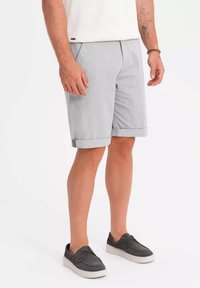 Shorts gris clair avec des revers, en tissu lisse. Portés avec des chaussures sombres à semelle blanche. Design décontracté.