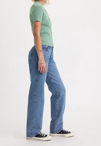 Green ribbed short-sleeve cardigan, light blue wide-leg jeans, and black sneakers with white soles. Visible tattoos on the forearm.