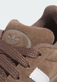 Brown suede sneaker featuring a textured surface, woven brown laces, and a white accent stripe, with a logo embossed on the tongue.