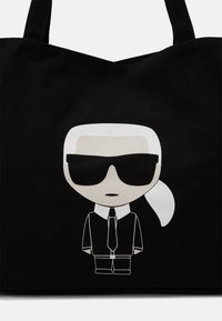 Malá stylizovaná postava s bílými vlasy staženými vzadu, černými slunečními brýlemi, černým kostýmem, bílou košilí a černou kravatou na černé tašce.