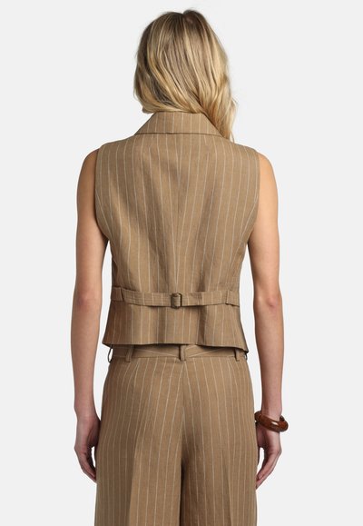 Szőke személy, aki ujjatlan, bézs csíkos mellényt visel hátul csattal, valamint hozzáillő csíkos nadrágot, háttal áll.