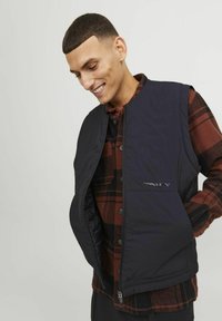 Zwarte gewatteerde vest met een rits aan de voorkant, gedragen over een rood-zwart geruite shirt. Bevat een logo en heeft een gladde textuur.