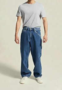 Homme portant un jean bleu ample, un t-shirt gris clair uni et des chaussures blanches, debout devant un fond neutre.
