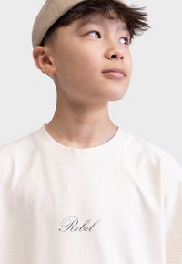 T-shirt en coton beige avec une coupe loose, un col rond et le mot "Rebel" imprimé en lettres cursives noires sur la poitrine.