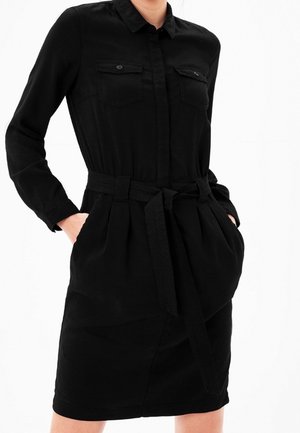 Mujer con un vestido negro de manga larga con botones, dos bolsillos en el pecho y un cinturón en la cintura, manos en los bolsillos laterales.