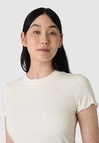 Úzký žebrovaný top s krátkým rukávem v krémové barvě, s kulatým výstřihem a jemně volánkovými manžetami. Hladká textura s těsným střihem.