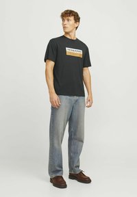 Sort bomulds T-shirt med en rektangulær grafik i hvid og orange med teksten "JACK & JONES." Matchet med lyseblå jeans og brune sko.