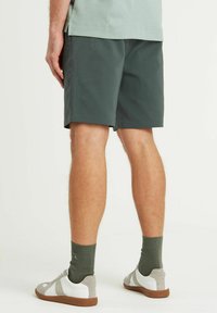 Pantaloni corti leggeri di color verde scuro con tasche laterali, caratterizzati da un design pulito. Indossati con scarpe grigie e bianche e calze verde a coste.