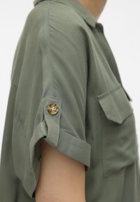 Camicia verde oliva a maniche corte realizzata in tessuto morbido, con un risvolto sul polsino dotato di un bottone dorato e una tasca sul petto.