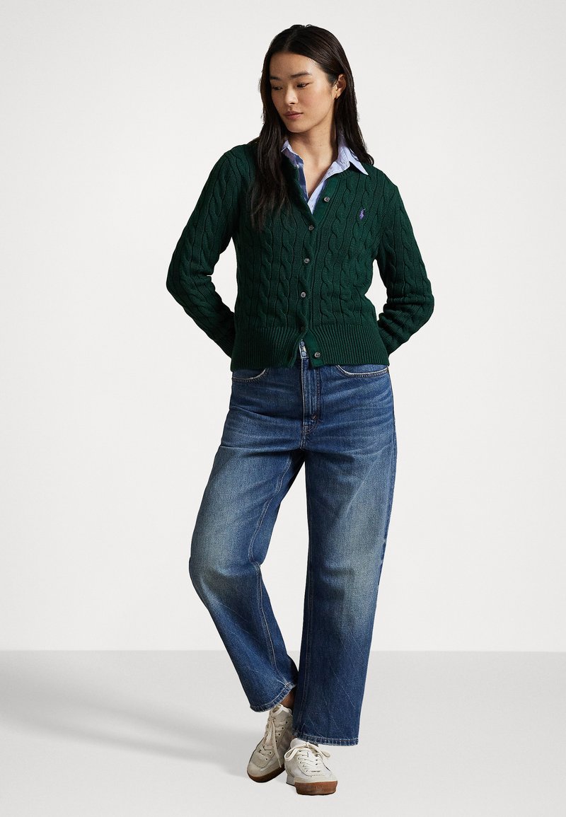 Grøn kabelstrikket cardigan med knaplukning, båret over en lyseblå skjorte med krave, parret med brede blå jeans og grå sneakers.