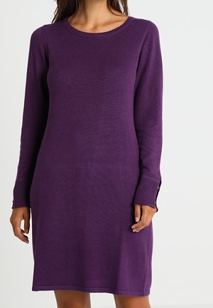 Vrouw die een paarse trui-jurk draagt met lange mouwen en een lengte tot aan de knie, voorzien van geribbelde manchetten en een ronde hals, tegen een effen achtergrond.