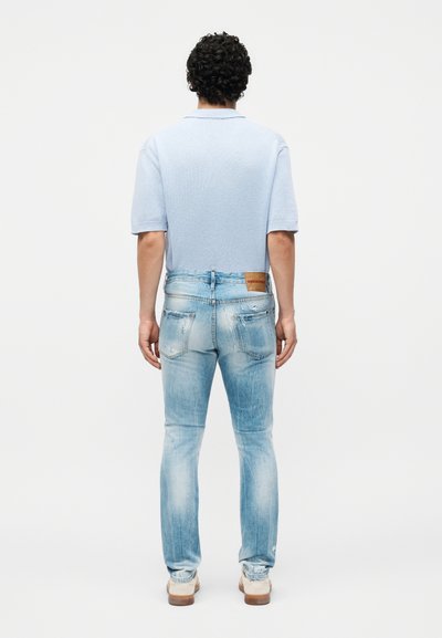 Άνδρας με σγουρά μαλλιά φορούν ανοιχτό μπλε κοντομάνικο πουλόβερ, ξεθωριασμένα μπλε τζιν με πίσω τσέπες και μπεζ αθλητικά παπούτσια, στέκεται γυρισμένος με την πλάτη.