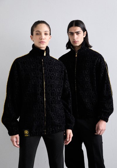 Zwarte zip-up jassen van een gestructureerde stof met herhaaldelijk "VERSACE" logo, gouden rits en geribbelde manchetten, gedragen met gestreepte zwarte broek.
