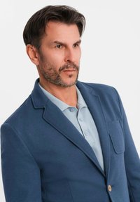 Man med skägg som bär en blå kavaj och en ljusblå polo t-shirt, tittar tankfullt åt sidan mot en enfärgad vit bakgrund.