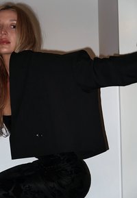 Svart kort blazer med överdimensionerade ärmar och en enda knappknäppning, bärs över en svart texturerad outfit, mot en neutral bakgrund.