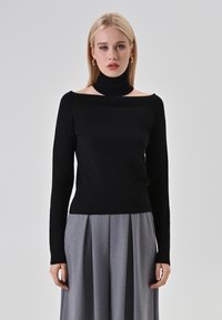 Maglione nero con colletto alto e design scendi spalle, realizzato in un tessuto morbido. Abbinato a pantaloni grigi a pieghe. Silhouette semplice e moderna.