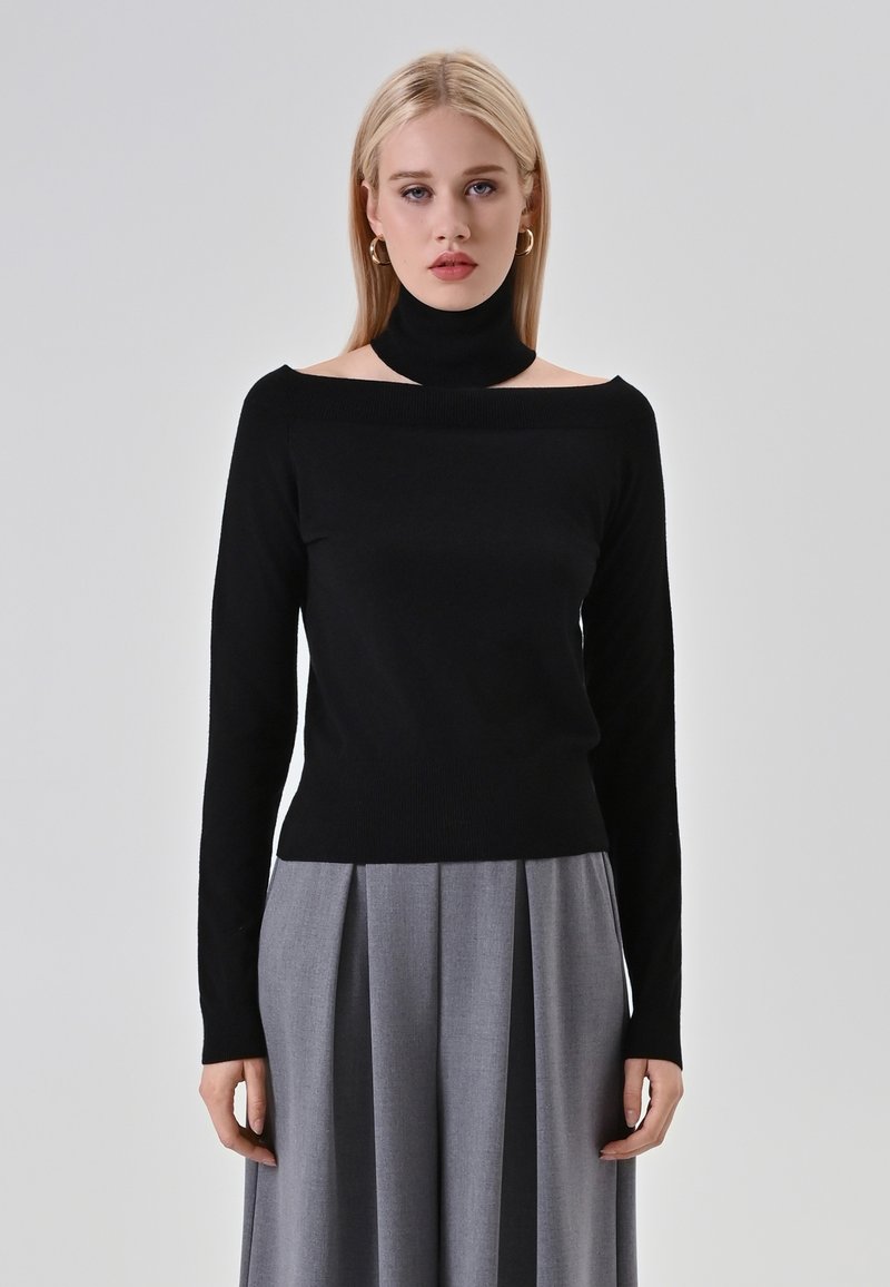 Maglione nero con colletto alto e design scendi spalle, realizzato in un tessuto morbido. Abbinato a pantaloni grigi a pieghe. Silhouette semplice e moderna.
