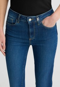 Jeans in denim blu scuro con una vestibilità slim, dotati di chiusura con bottone, design a cinque tasche e cuciture a contrasto. Texture liscia, dettagli minimi.
