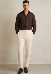 Camisa de lino marrón de botón con mangas largas, pantalones cremosos ajustados y zapatos oscuros de tipo slip-on. Diseño sencillo, silueta ajustada.
