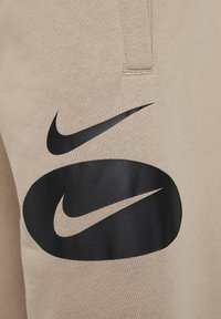 Béžové tepláky s jemnou texturou, zdobené velkým černým logem Nike a menším motivem swoosh. Dokončeno s běžným pasem.