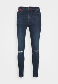 Vaqueros ajustados de mezclilla azul oscuro, con una cintura alta, rasgaduras en las rodillas y un sutil patrón de lavado.