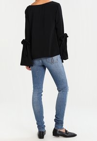 Top negro de manga larga con puños anchos atados en las muñecas, combinado con jeans ajustados azules y zapatos planos negros, visto desde atrás.