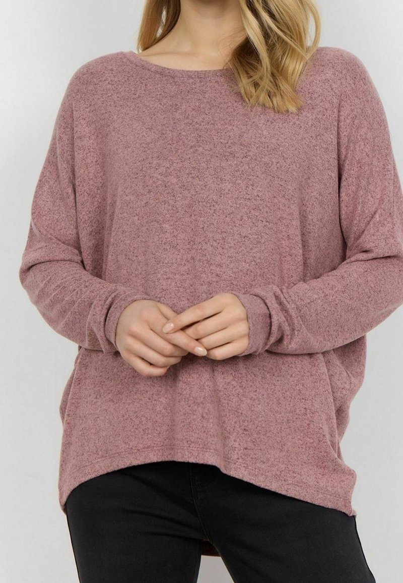 Femme portant un pull ample à manches longues de couleur mauve et un pantalon noir, les mains jointes à la taille, sur un fond clair uni.
