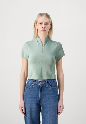Mint green short-sleeve crop top with a stand collar and front zipper, made of smooth fabric, paired with high-waisted blue jeans.