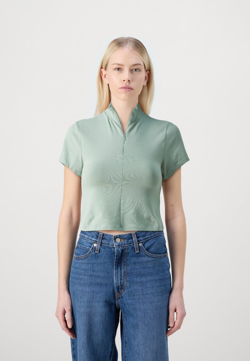 Top corto de manga corta en verde menta con cuello alto y cremallera frontal, hecho de tejido suave, combinado con unos vaqueros de cintura alta en azul.