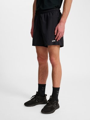Mann trägt schwarze Sportshorts mit Reißverschlusstasche, schwarze Kniestrümpfe und schwarze Laufschuhe vor weißem Hintergrund.