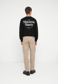Hanorac negru cu textul "Nineteen Ninety" pe spate, asortat cu pantaloni cargo bej cu buzunare și adidași negri groși.