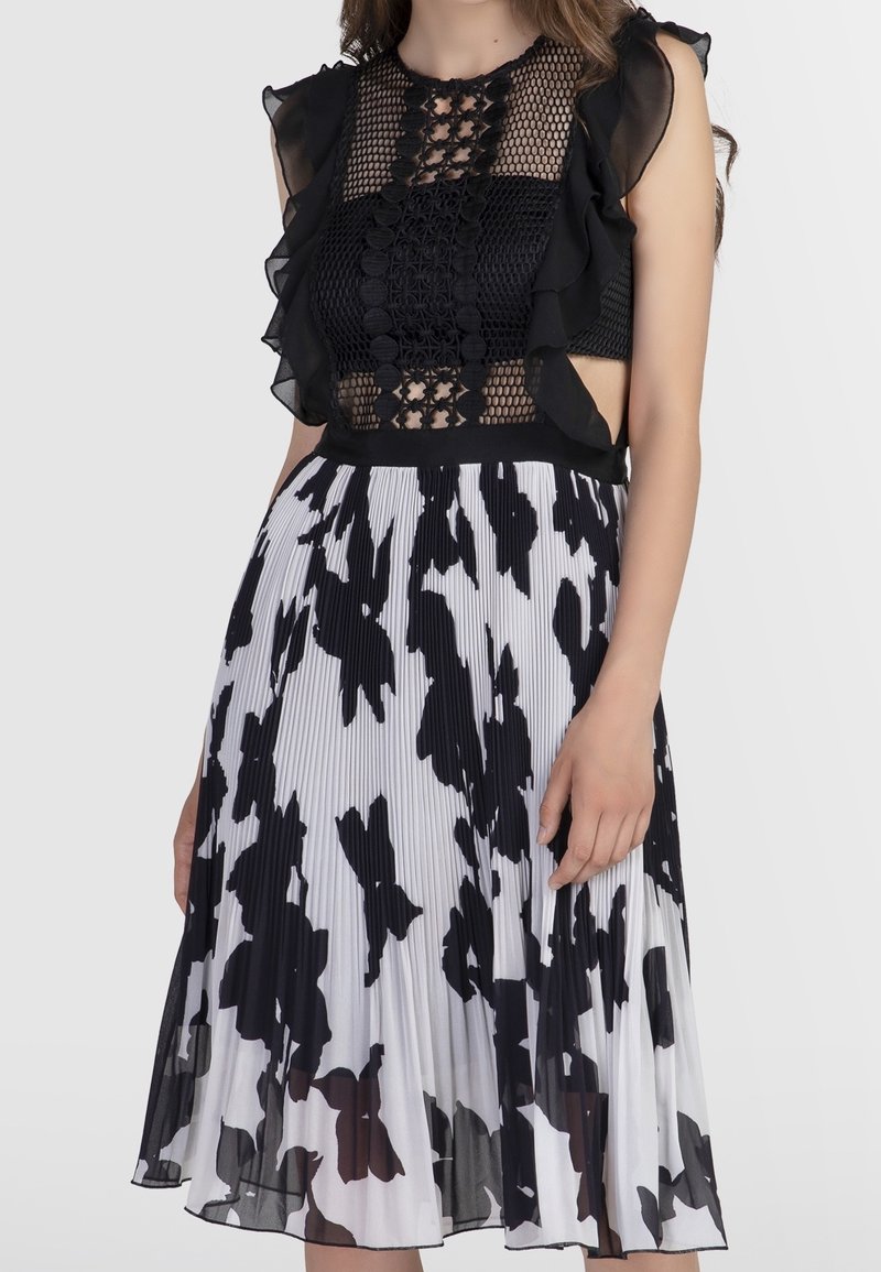 Femme portant un haut sans manches en dentelle noire avec des volants et une jupe plissée blanche avec un imprimé floral abstrait noir audacieux.