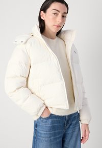 Puffjakke, hvit, kort design med høy krage, frontglidelås og elastiske mansjetter. Brukt over en kremfarget genser og denim.