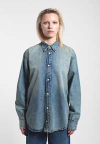 Camicia di denim con un ampio colletto, chiusura frontale con bottoni e maniche lunghe. Gradiente blu chiaro a blu scuro con texture sottile e dettagli di cucitura.