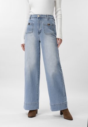 Žena oblečená v svetlomodrých džínsach s širokými nohavicami, prednými našitými vreckami a hnedými čižmami na podpätku, v kombinácii s bielym rebrovaným dlhým rukávom.