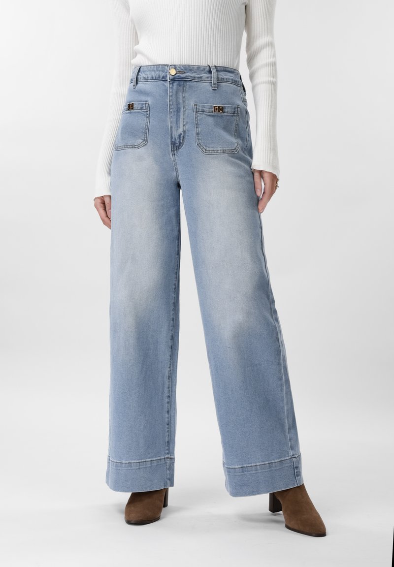 Γυναίκα που φοράει ανοιχτό μπλε τζιν παντελόνι με πλατιά μπατζάκια και μπροστινές τσέπες, και καφέ μποτάκια με τακούνι, σε συνδυασμό με ένα λευκό ριμπ μπλουζάκι μακρυμάνικο.