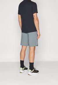 Mann in schwarzem Poloshirt, grauen Shorts, schwarzen Socken und schwarz-weißen Sneakers, der mit dem Rücken zur Kamera vor einem einfarbigen Hintergrund steht.