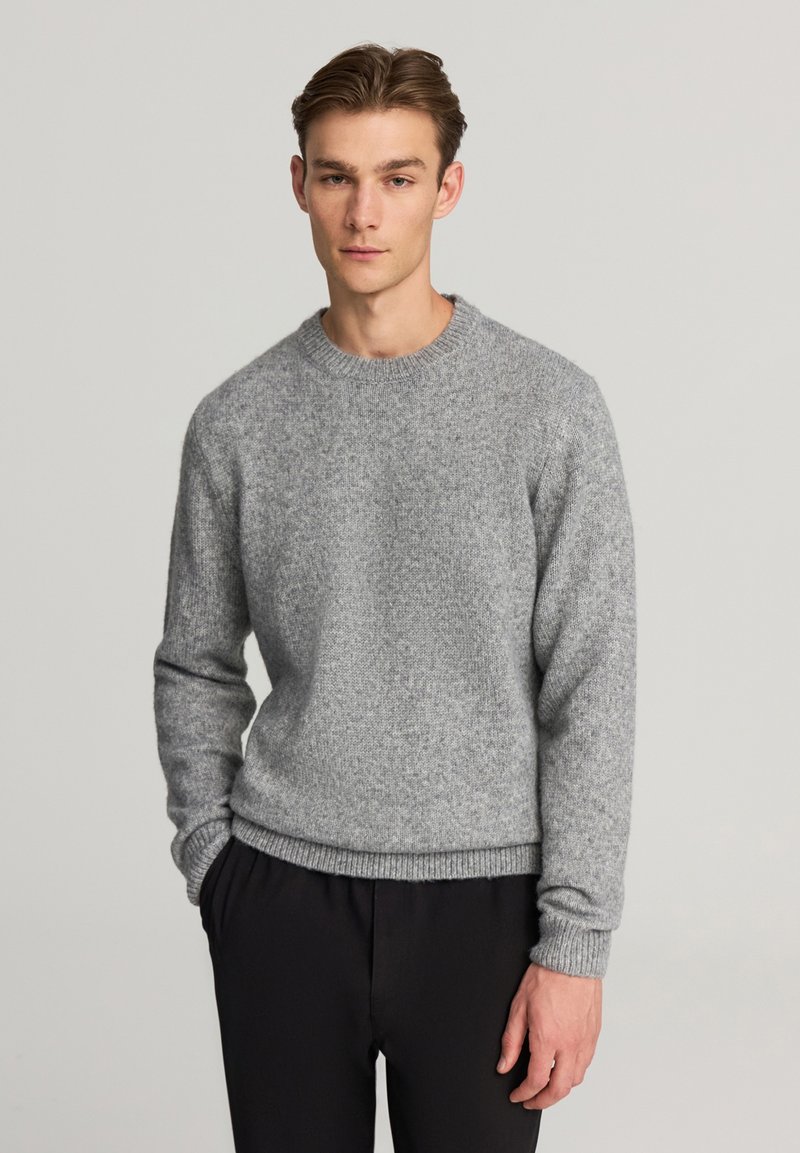 Maglione grigio lavorato a maglia realizzato in tessuto testurizzato. Presenta un colletto rotondo, polsini e orlo a coste. Indossato con pantaloni neri.