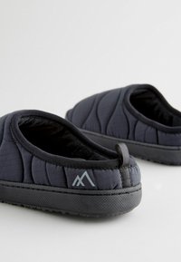 Pantofole trapuntate nere con suola testurizzata, lati imbottiti e una linguetta. Il design include un piccolo logo di una montagna grigia sul lato.