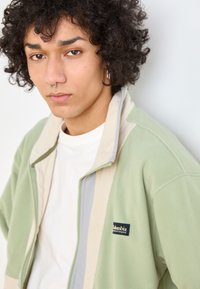 Giacca in fleece verde chiaro con accenti beige e grigi, caratterizzata da una chiusura con zip e un'etichetta del logo Columbia sul lato sinistro. Tessuto morbido.