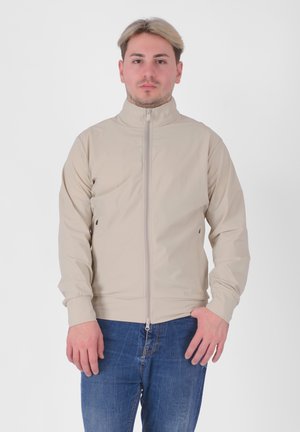 Giacca beige con colletto alto, zip frontale completa e polsini elasticizzati; presenta un design minimalista e una texture liscia. Indossata sopra jeans blu.