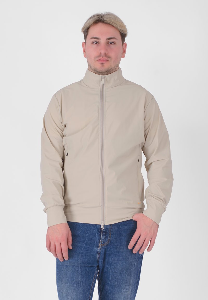 Giacca beige con colletto alto, zip frontale completa e polsini elasticizzati; presenta un design minimalista e una texture liscia. Indossata sopra jeans blu.