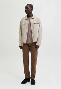 Casaco de ganga bege com dois bolsos frontais, t-shirt cinzenta, calças castanhas e sapatos pretos; modelo está em pé com uma expressão neutra contra um fundo simples.