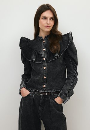 Vrouw met lang bruin haar die een zwarte spijkerjas draagt met geplooide schouders en een bijpassende zwarte spijkerbroek, handen in de zakken.