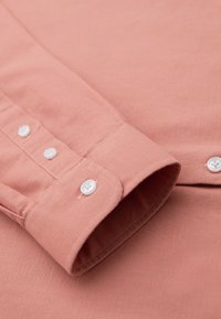 Gros plan sur un poignet de chemise à manches longues couleur saumon avec trois boutons blancs et une texture de tissu visible.
