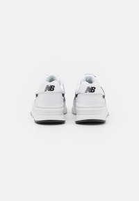 Pár bílých tenisek New Balance s černým logem na patách, z pohledu zezadu na bílém pozadí.